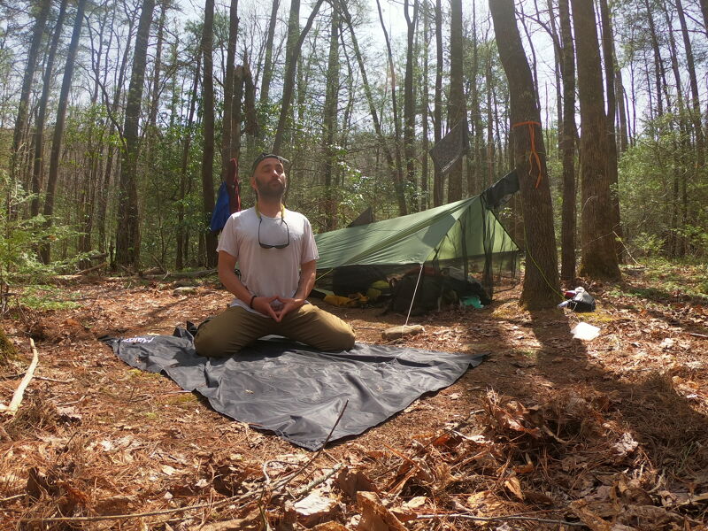 A man is meditating on a black mat in a forest clearing. He is kneeling with his hands in a prayer position, eyes closed, and head tilted upwards. He is wearing a white t-shirt and khaki pants. Behind him, there is a green tent set up among the trees. The ground is covered with leaves and pine needles, and the sun is shining through the trees.